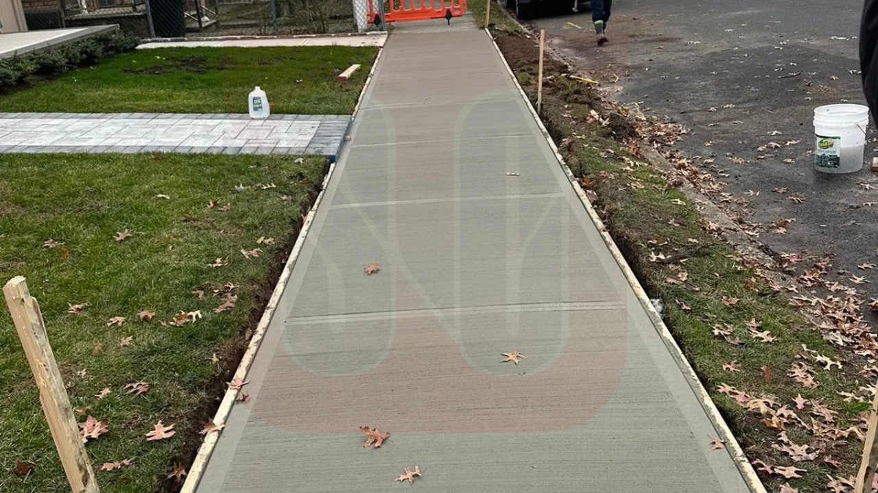 A freshly poured concrete sidewalk with scattered leaves, bordered by grass and a road, with construction materials and a worker in the background
